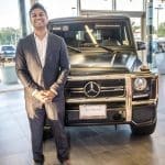 A smiling man in a gray suit stands beside a matte black Mercedes-Benz G-Class SUV in a showroom.