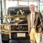 A smiling man in a brown blazer stands beside a matte black Mercedes-Benz G-Class SUV in a showroom.