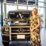 A woman in a floral jumpsuit stands beside a matte black Mercedes-Benz G-Class SUV inside a showroom.