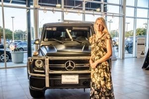 A woman in a floral jumpsuit stands beside a matte black Mercedes-Benz G-Class SUV inside a showroom.