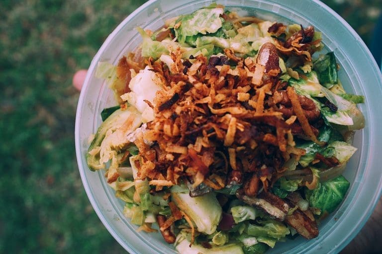 A plastic bowl of shredded green salad topped with crispy fried onion strings, pecans, and dressing, held outdoors over grass.