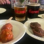 Two paper plates hold saucy meatballs and chocolate dessert bites beside two beers in glasses printed "flavor.