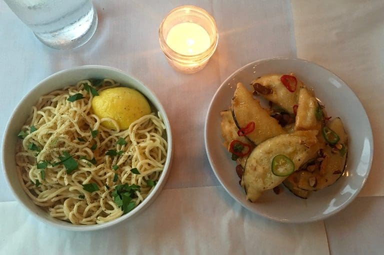 Overhead view of spaghetti with parsley and a lemon half beside fried zucchini with chili slices and almonds, next to a candle.