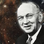 Black-and-white portrait of a smiling older man in wire-rimmed glasses and suit, set against a starry nebula background.