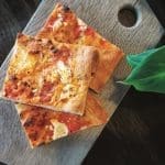 Three square slices of tomato and cheese flatbread on a wooden board, with a green leaf beside them.