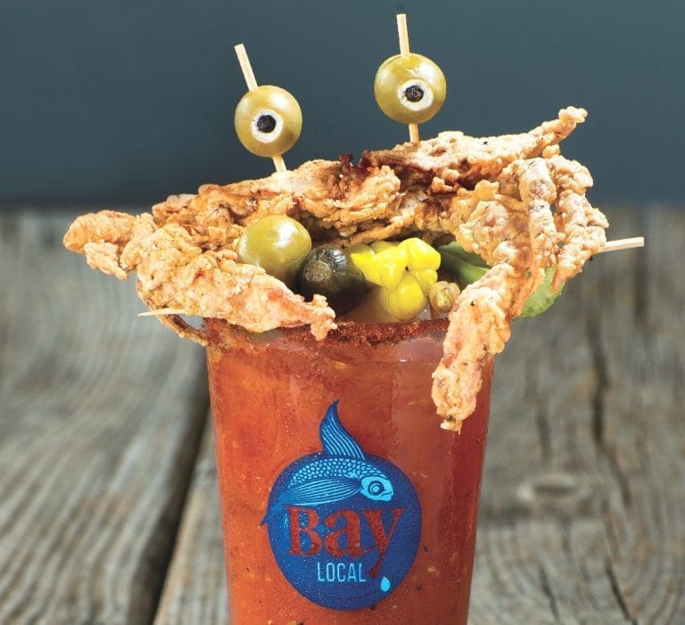 A Bloody Mary in a Bay Local glass topped with a fried soft-shell crab, olives on picks, and pickled vegetables.