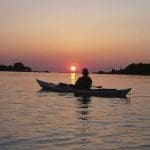 A silhouetted kayaker paddles across calm water toward a setting sun over distant tree-lined shoreline.