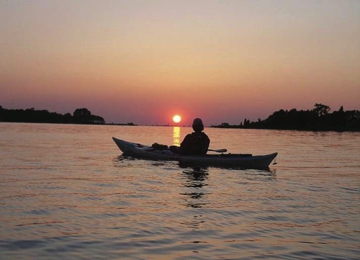 A silhouetted kayaker paddles across calm water toward a setting sun over distant tree-lined shoreline.