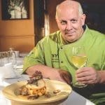 Smiling chef in a lime-green jacket holds white wine beside a plated fish dish with crispy garnish and roasted vegetables.