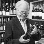 Smiling older man in glasses and a plaid blazer holds a red wine bottle in a wine shop.