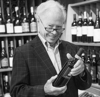 Smiling older man in glasses and a plaid blazer holds a red wine bottle in a wine shop.