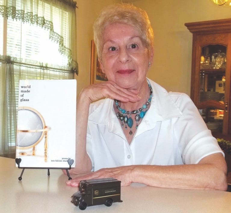 Smiling woman with short blonde hair and turquoise necklace beside a "world made of glass" book on an easel and a toy brown truck.