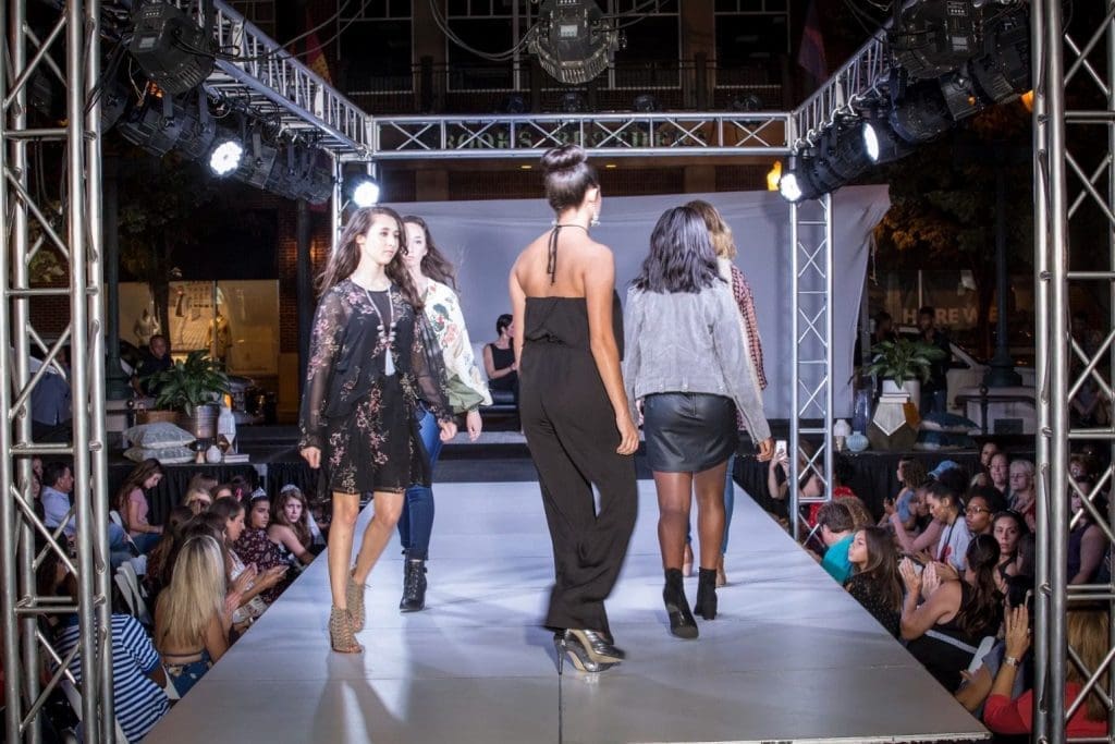 Models walk a runway lined with spectators under stage lights during an evening fashion show.