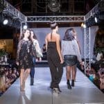 Models walk a runway lined with spectators under stage lights during an evening fashion show.