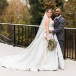 A bride in a white satin ballgown with a long veil and a groom in a gray suit with purple boutonniere pose on a deck.