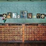 A long wooden front desk made of rows of small apothecary-style drawers, with framed portraits and a teal patterned wallpaper behind it.