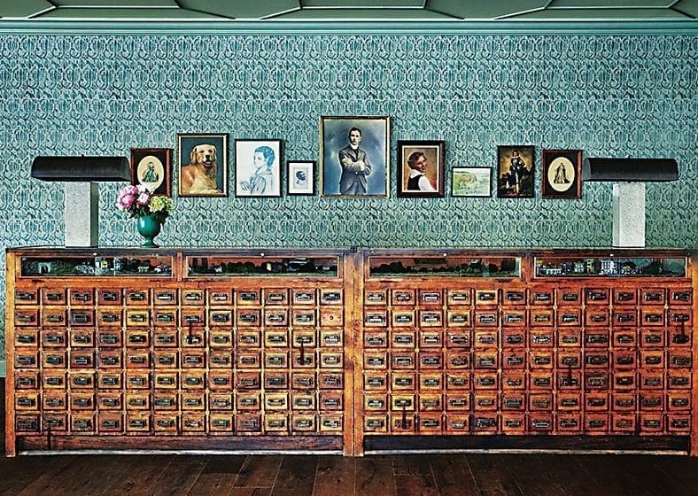 A long wooden front desk made of rows of small apothecary-style drawers, with framed portraits and a teal patterned wallpaper behind it.