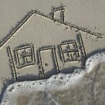 House in the sand is overtaken by a foamy wave; focus on sand and motion blur on the wave
