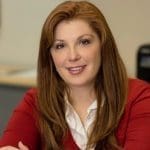 Smiling woman with long auburn hair in a red cardigan over a white collared shirt, seated in an office.