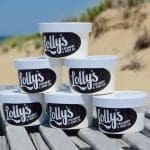 Stacked white pint containers labeled "Lolly's Creamery & Soup Co." sit on a weathered wooden boardwalk with sand dunes behind.