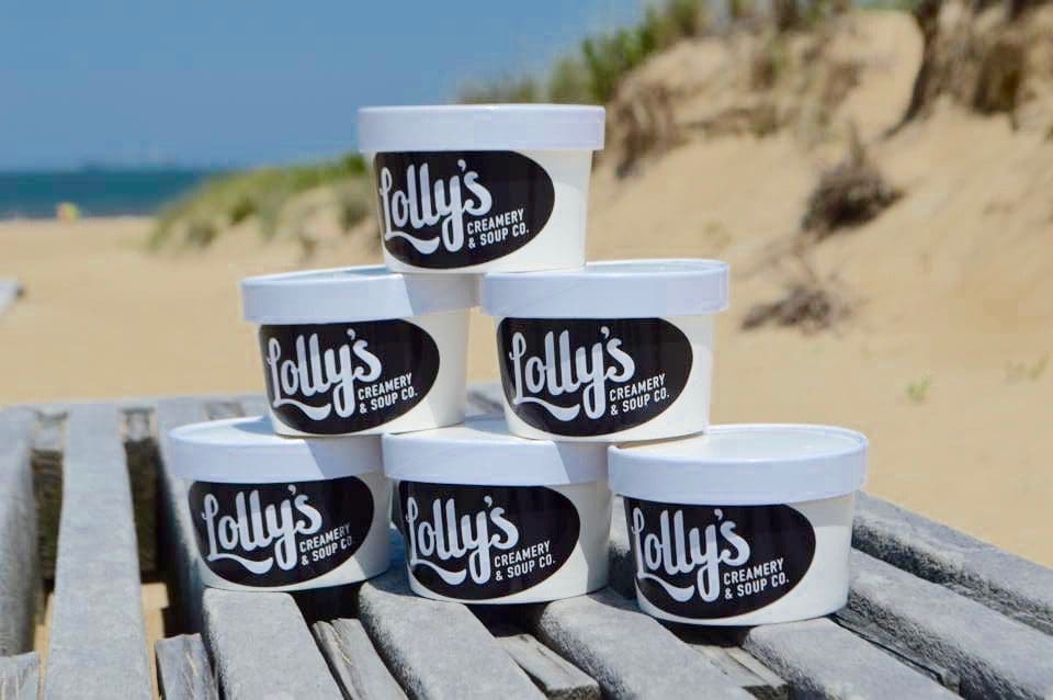 Stacked white pint containers labeled "Lolly's Creamery & Soup Co." sit on a weathered wooden boardwalk with sand dunes behind.