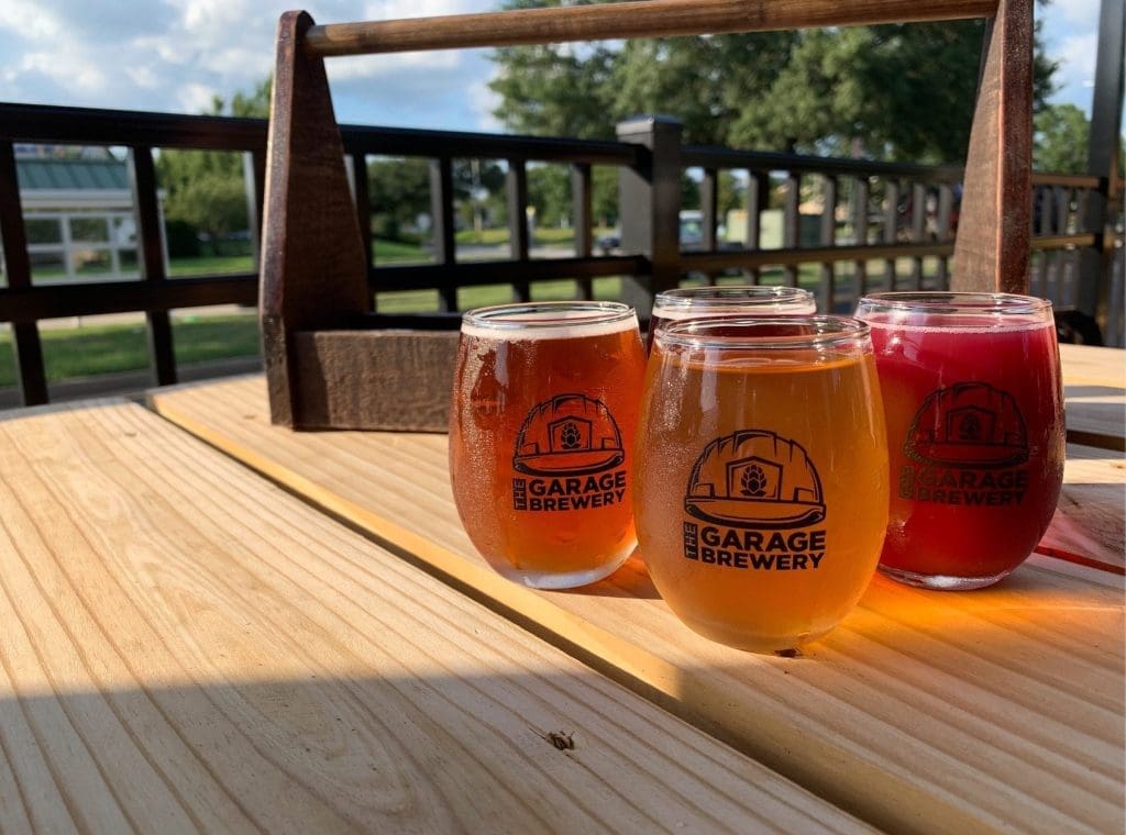 Four stemmed sampler glasses of amber, golden and pink beer, branded "The Garage Brewery," on a wooden tray outdoors.