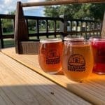 Four stemmed sampler glasses of amber, golden and pink beer, branded "The Garage Brewery," on a wooden tray outdoors.