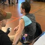 A tattoo artist in a gloved hand works on a seated man's upper arm, a stencil drawn on his skin, in a gym.