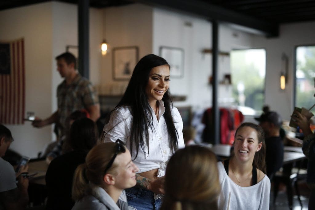 A smiling woman with long dark hair in a white shirt stands among seated patrons in a bright café.