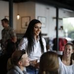 A smiling woman with long dark hair in a white shirt stands among seated patrons in a bright café.