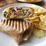 Grilled wrap cut in half showing sliced turkey, avocado, bacon and tomato, served with potato chips on a white plate.