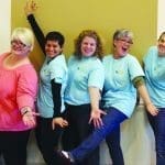 Five smiling women in light blue t-shirts pose playfully against a wall, arms and legs akimbo.