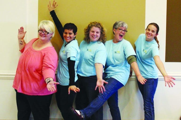 Five smiling women in light blue t-shirts pose playfully against a wall, arms and legs akimbo.
