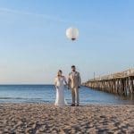 A bride in a white gown and a groom in a tan suit hold hands on a sandy beach beside a long wooden pier, a large white balloon overhead.