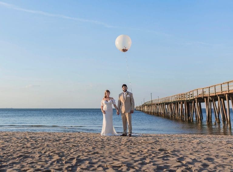 A bride in a white gown and a groom in a tan suit hold hands on a sandy beach beside a long wooden pier, a large white balloon overhead.