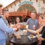 Six people clink plastic beer cups in front of the Festhaus, a wooden Bavarian-style building with a sign, at an outdoor table.