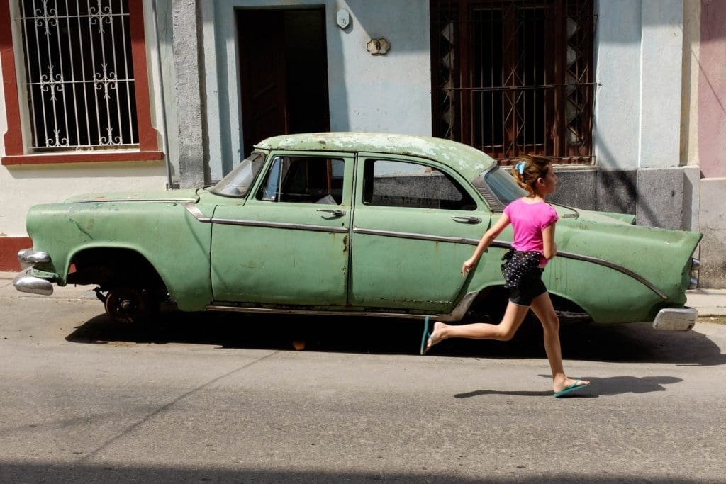 A girl in a pink shirt runs past a weathered green vintage car on a sunlit Havana street.