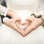 A bride in white lace and a groom in a dark suit form a heart with their hands at her waist.