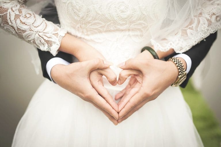 A bride in white lace and a groom in a dark suit form a heart with their hands at her waist.