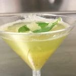 A pale yellow-green cocktail in a clear martini glass, garnished with fresh basil leaves and shaved ice or frost.