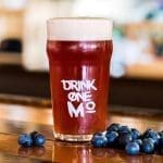 A pint glass of reddish-amber beer with white foam, printed "DRINK ONE MO", beside fresh blueberries on a wooden bar.