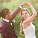 A bride in a lace strapless gown twirls under her raised arm beside a groom in a plum suit, outdoors among green trees.