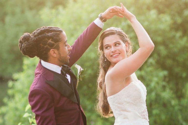 A bride in a lace strapless gown twirls under her raised arm beside a groom in a plum suit, outdoors among green trees.
