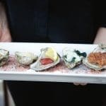 A server in black holds a white platter of five dressed oysters on pink salt, one topped with lemon.