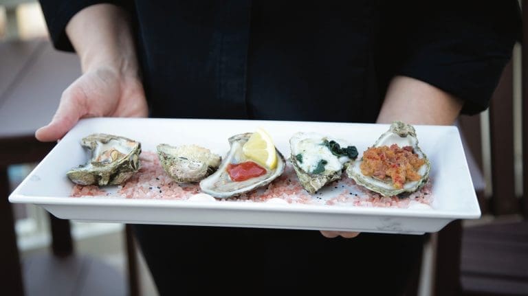 A server in black holds a white platter of five dressed oysters on pink salt, one topped with lemon.