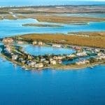 Aerial view of a waterfront neighborhood of homes and docks on a peninsula, with marshland, causeway bridge, and blue bay water.