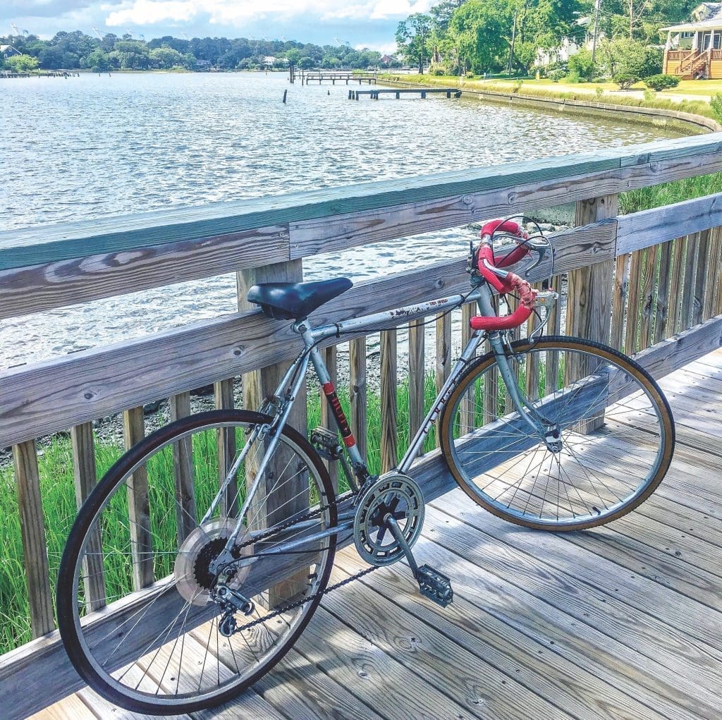 A silver road bike with red handlebar tape leans against a wooden railing on a waterfront boardwalk.