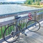 A silver road bike with red handlebar tape leans against a wooden railing on a waterfront boardwalk.