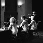 Black-and-white view of three seated panelists at a microphone-lined table as a speaker stands at a lectern.