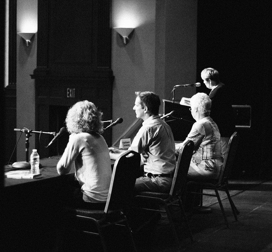 Black-and-white view of three seated panelists at a microphone-lined table as a speaker stands at a lectern.